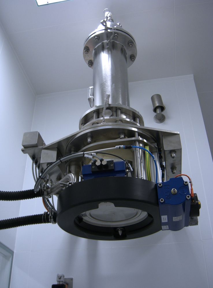Bottom-up view of a HECHT vibrating dosing valve mounted on the ceiling, featuring a cylindrical stainless steel body with a motor and control units attached. This valve is integral to controlling material flow in processing systems, designed for efficiency and cleanliness in high-purity environments.