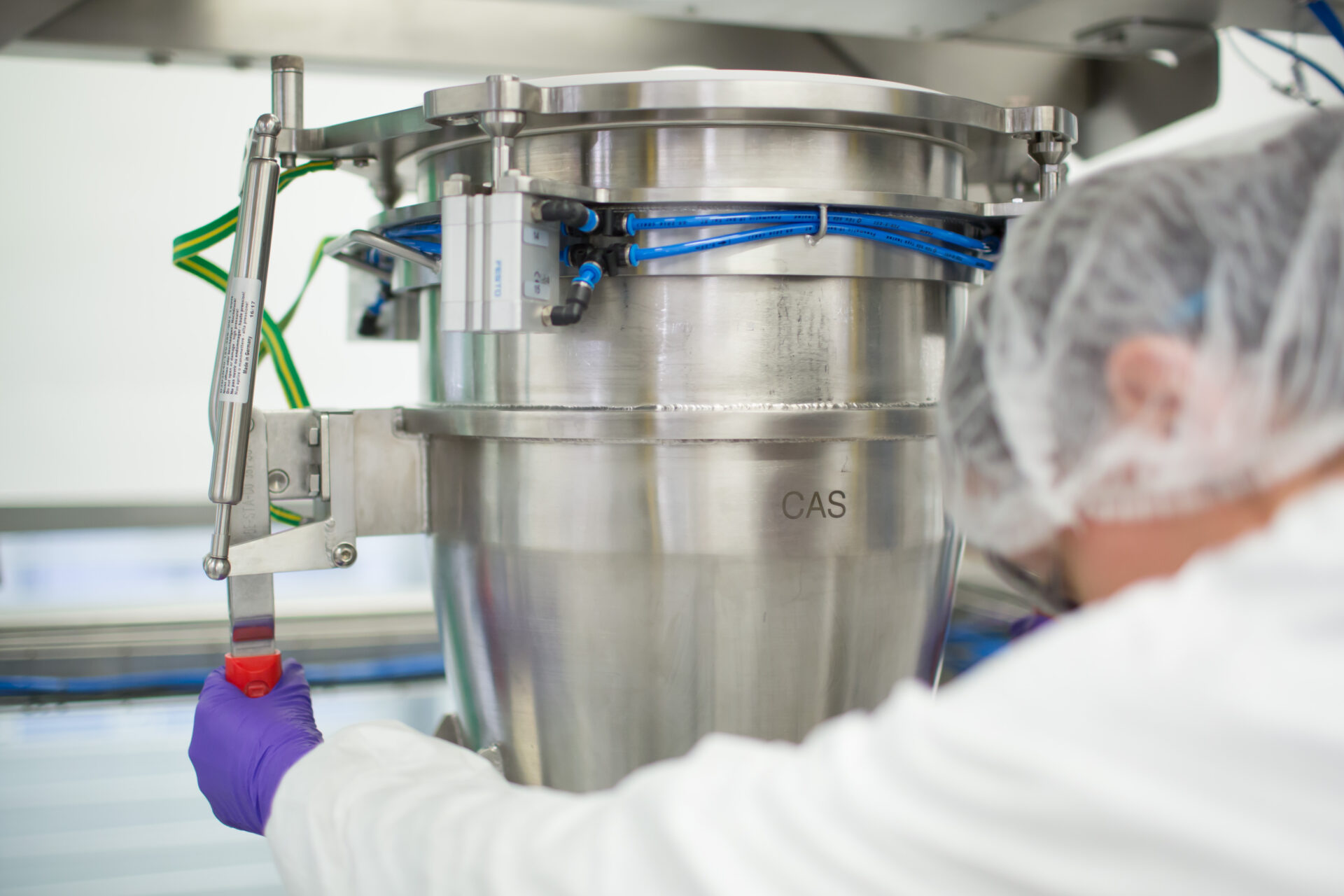 A technician in a cleanroom suit operates a HECHT Big Bag discharge station, focusing on the control mechanism with precision, set against the backdrop of a sterile, high-grade industrial environment, indicating stringent health and safety standards in material handling.