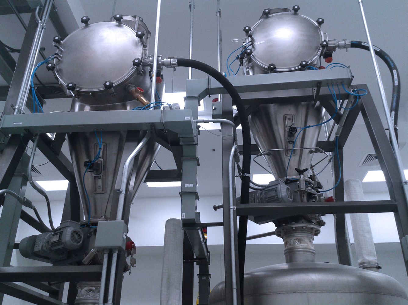 A pair of PCC 700 machines in a food production facility with stainless steel conical hoppers mounted on metal frames. The equipment, featuring a series of clamps, valves, and flexible tubing, is designed for bulk material handling in a hygienic environment.