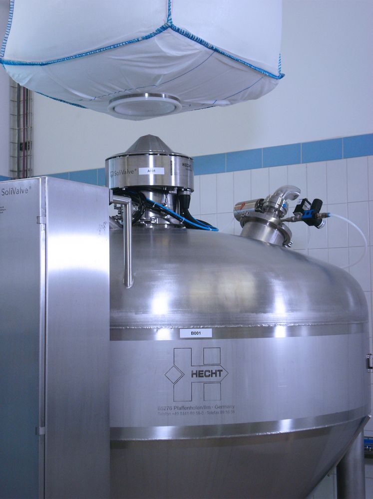 A HECHT BigBag discharge station with the SoliValve cone system, featuring a large suspended BigBag above a stainless steel funnel with a valve system, designed for precise, contained material transfer in a clean, tiled industrial setting.