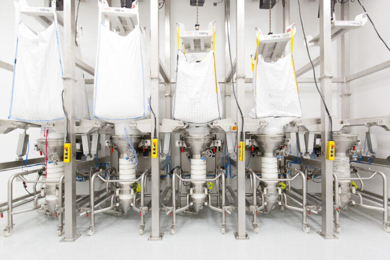 Industrial bulk material handling facility featuring HECHT equipment, with multiple white Big Bags suspended above stainless steel funnels for material discharge, all part of a meticulously organized system designed for high-capacity and efficient processing.