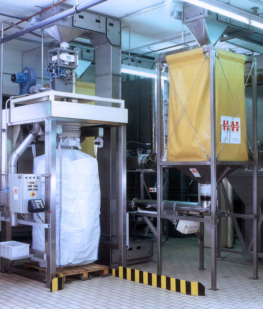 Industrial HECHT bag filling station, showcasing a white bulk bag being filled, with a yellow dust containment system overhead, set within a facility with metal structures and ductwork, emphasizing cleanliness and precision in bulk handling processes.