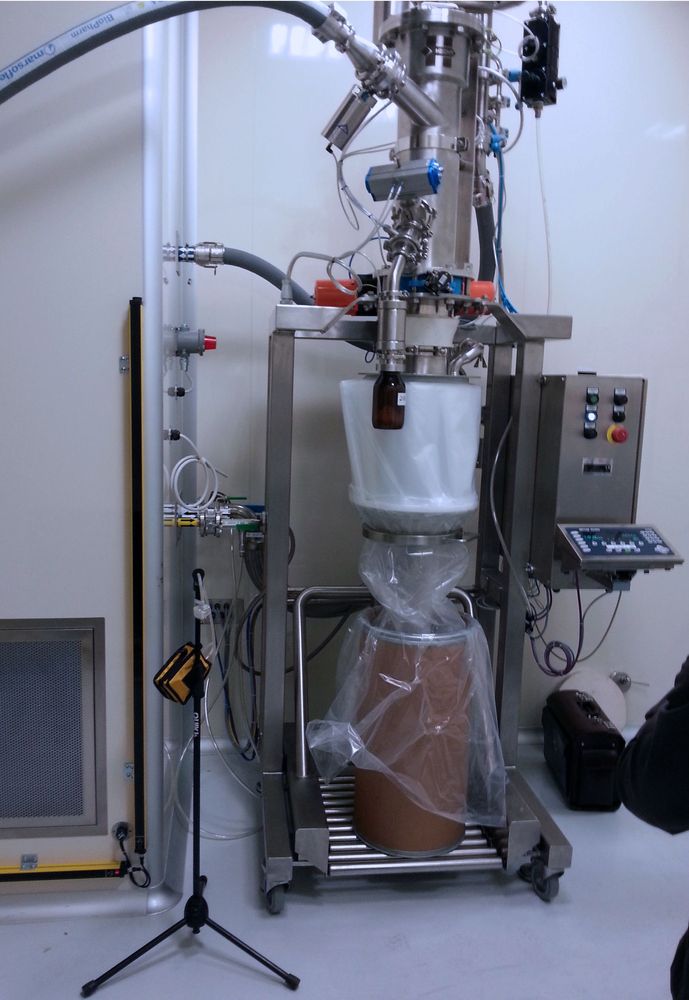 Alt text: "HECHT vacuum sampler at a small packaging station, with a stainless steel structure and a glass collection bottle, filling a white fiber drum inside a clear plastic bag, complemented by control panels and flexible tubing, in a controlled industrial setting.