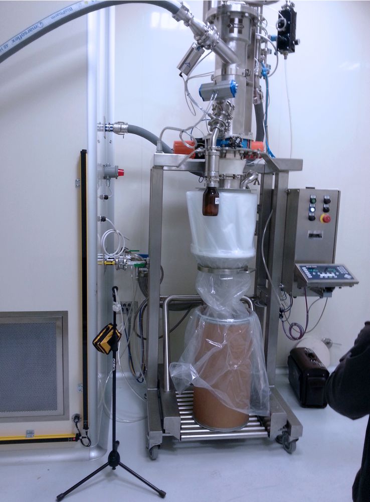 HECHT ProClean Conveyor (PCC) 200 system configured for drum filling and weighing operations, featuring a stainless steel funnel atop a cylindrical vessel that dispenses into a drum within a protective plastic sleeve. The setup includes a digital control panel on the right, with various cables and a portable scale in the foreground.