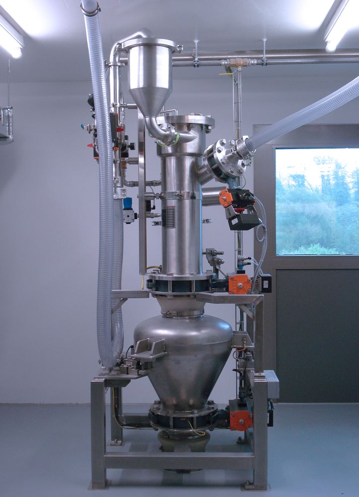Vertical setup of a HECHT ProClean Conveyor (PCC) 200 system with WIP unit, consisting of a stainless steel hopper, connecting flexible hoses, and a conical bottom processing vessel. The system is mounted on a metal frame within a clean room with visible piping in the background and a window showing a natural outdoor view.