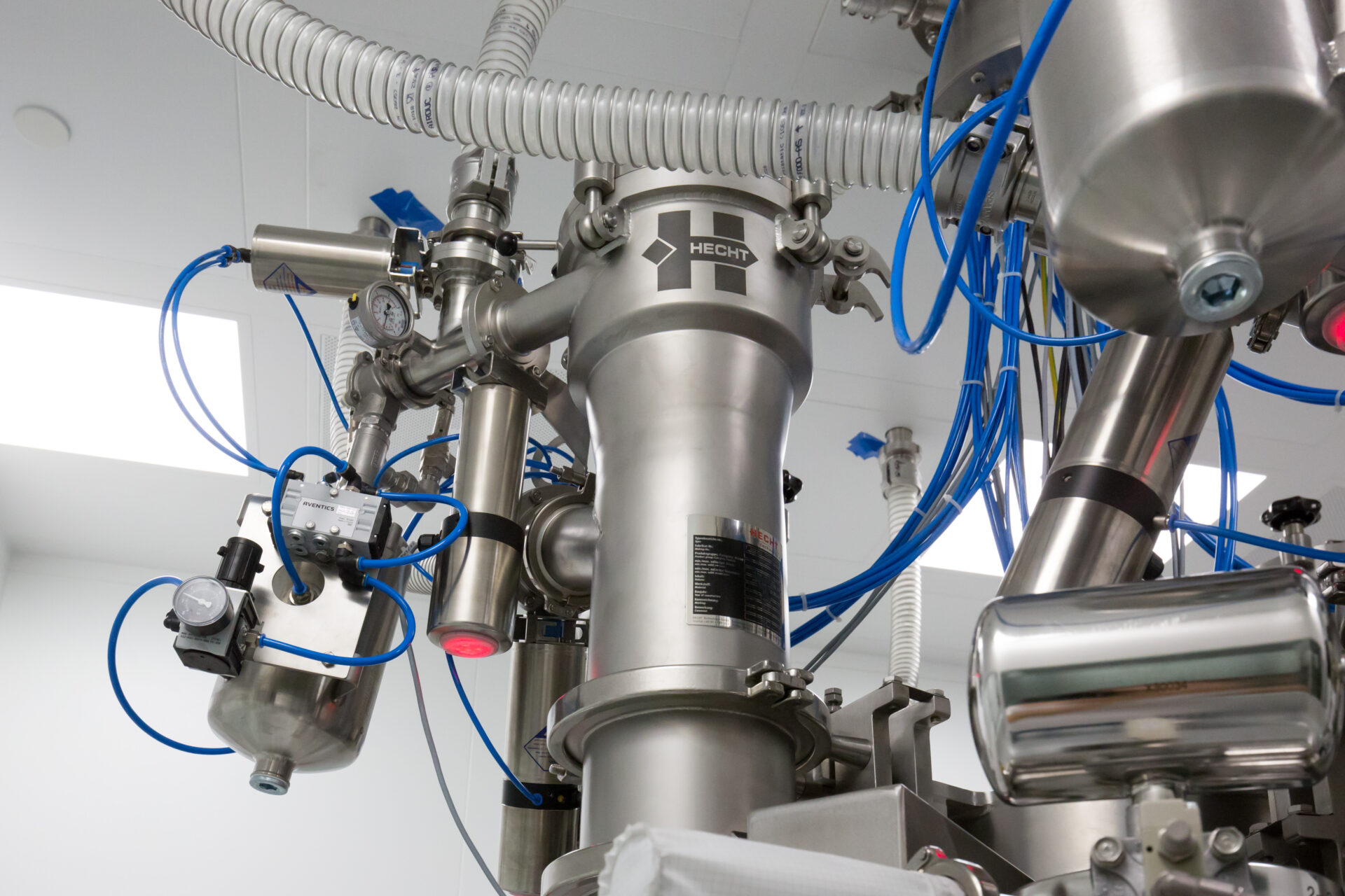 Complex HECHT ProClean Conveyor machinery at Pfizer Germany, featuring stainless steel components, tubing, pressure gauges, and flexible conduits. The system's intricate design and precise instrumentation are set against a clean, white ceiling with industrial lighting.